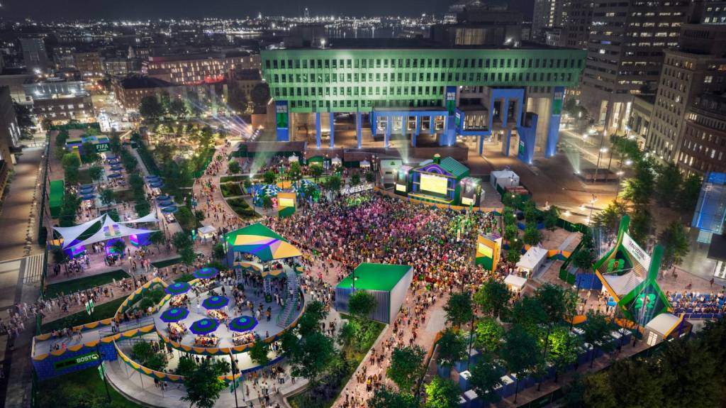Vista aérea noturna de um festival ao ar livre com multidão, palcos iluminados e tendas coloridas. Um grande edifício verde e azul se destaca ao fundo, enquanto luzes de cidades e árvores completam o cenário festivo