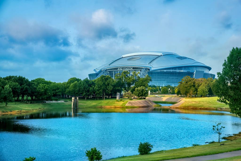 Estádio AT&T em Arlington, Texas, com sua cúpula prateada e fachada de vidro, visto por trás de um lago azul e árvores verdes sob um céu parcialmente nublado