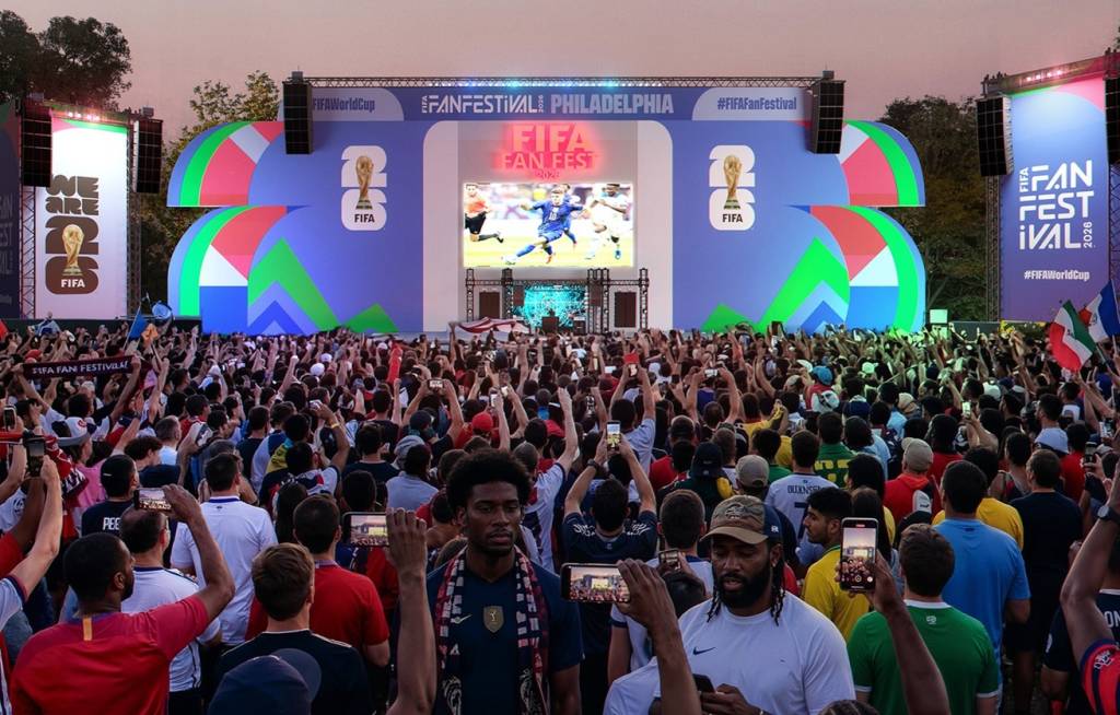 Multidão assiste a um jogo de futebol em telão gigante do FIFA Fan Fest Philadelphia 2026, com pessoas filmando e erguendo os braços. O palco tem a taça da Copa do Mundo e o logo da FIFA