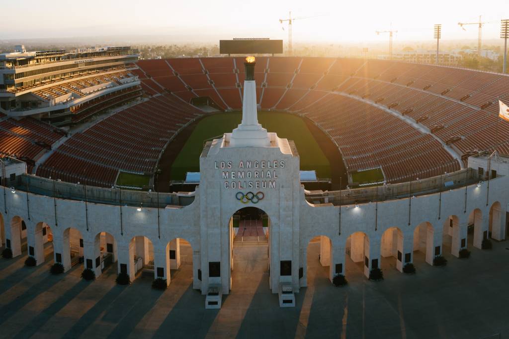 Vista aérea do Los Angeles Memorial Coliseum ao pôr do sol, com a tocha olímpica acesa no topo da entrada principal. O estádio tem arquibancadas vermelhas vazias e um campo verde. A fachada de pedra clara exibe