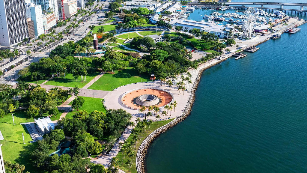 Vista aérea de um parque verdejante com palmeiras e um monumento circular de pedra, margeando um corpo d'água azul-esverdeado. Ao fundo, prédios altos, ruas movimentadas e uma marina com barcos e uma roda-gigante.