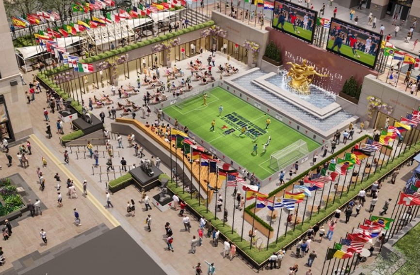 Vista aérea de uma fan fest de futebol no Rockefeller Center, com um campo de futebol verde, pessoas jogando e assistindo, e bandeiras de vários países alinhadas em passarelas. Duas telas grandes exibem partidas, e uma fonte dourada com estátuas adorna o centro do local. Muitas pessoas circulam e relaxam em cadeiras de praia