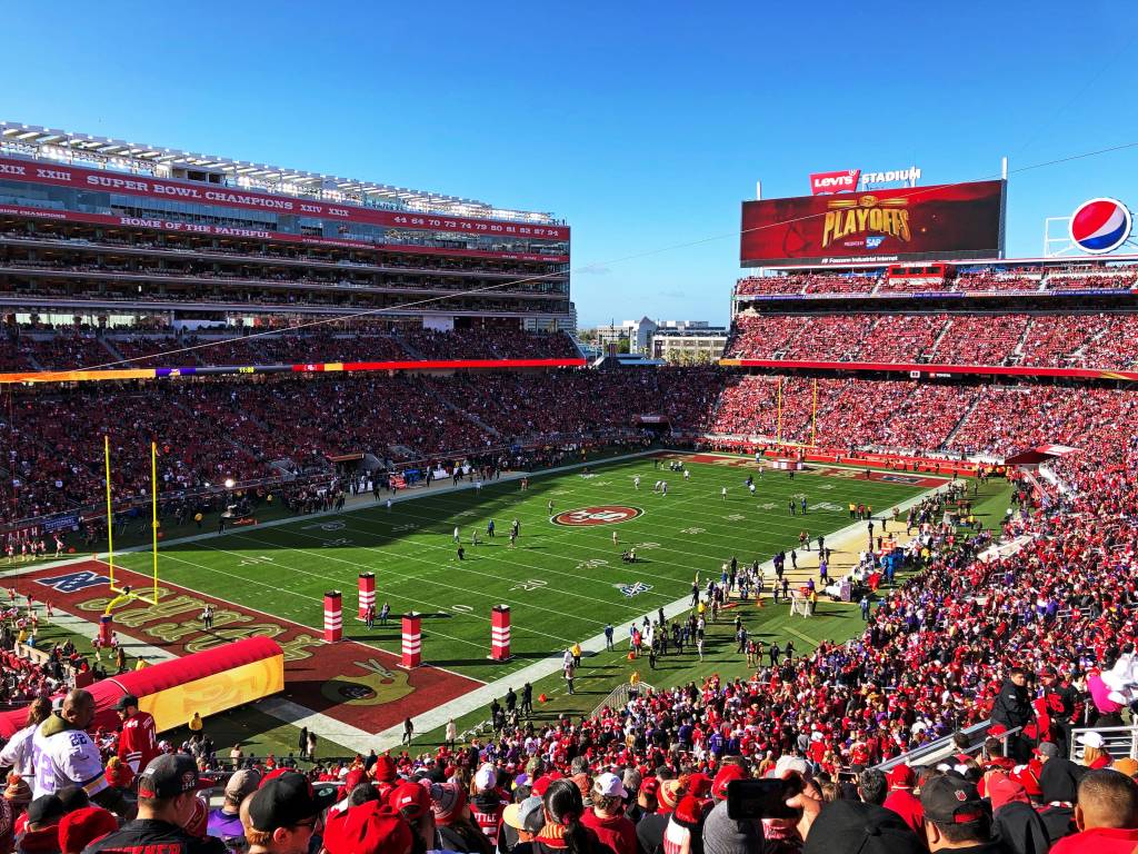 Vista aérea de um estádio de futebol americano lotado, com torcedores vestindo vermelho. O campo verde tem marcações brancas e jogadores em campo. Telões exibem