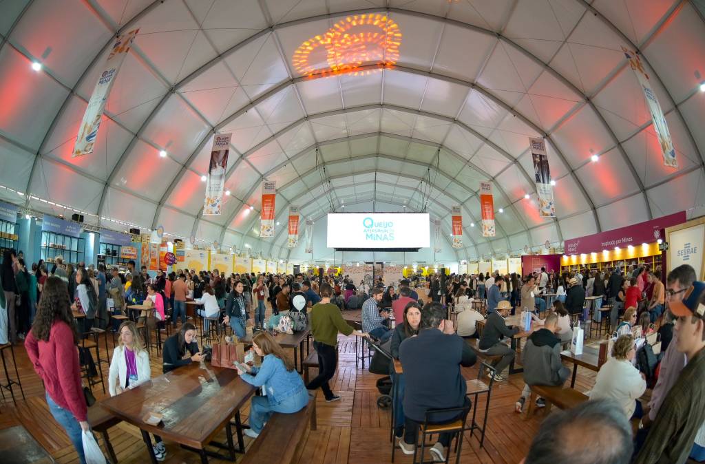 Grande tenda branca com centenas de pessoas em um evento, algumas sentadas em mesas de madeira, outras circulando. Um telão exibe