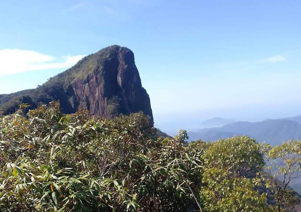 Montanha rochosa e íngreme, coberta por vegetação verde no topo, sob céu azul claro. Em primeiro plano, arbustos e folhagens densas. Ao fundo, outras montanhas e um vale com névoa.