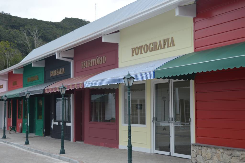 Fachadas coloridas de lojas com toldos listrados e lisos, incluindo uma barbearia, escritório e fotografia, em uma rua com postes de luz e montanhas ao fundo