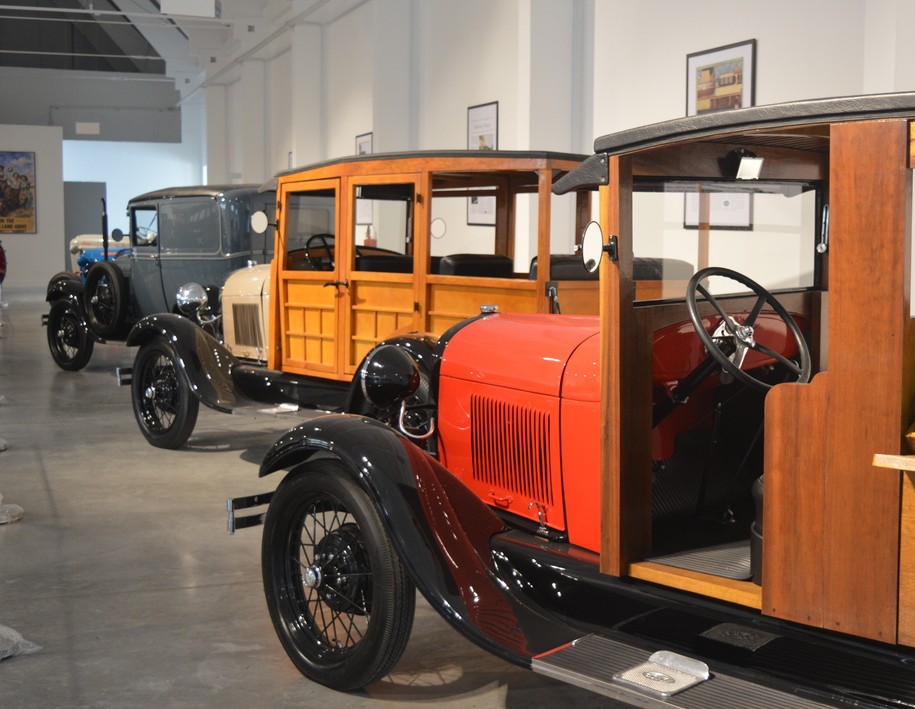 Três carros antigos em exposição, o primeiro vermelho e preto com carroceria de madeira, o segundo bege e madeira, e o terceiro azul-acinzentado, todos com pneus pretos e aros finos, em um salão com paredes brancas e piso cinza