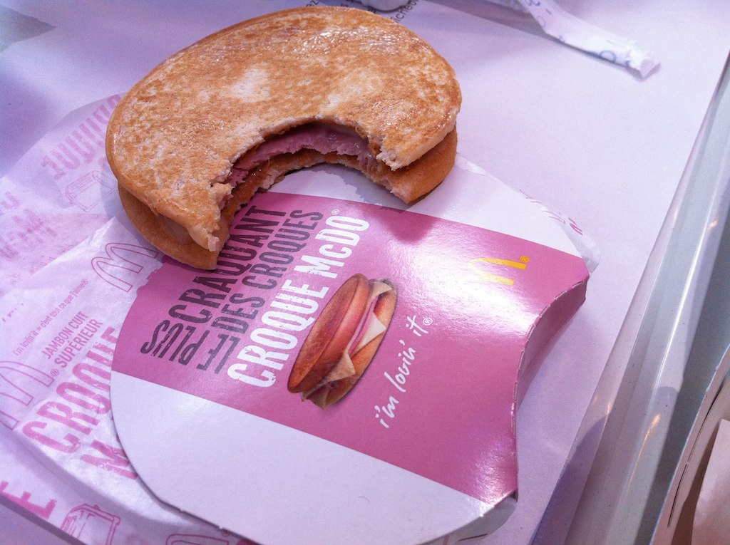 Sanduíche Croque McDo mordido, mostrando o recheio de presunto e queijo, sobre embalagem rosa e papel de embrulho do McDonald's, em uma bandeja branca. Foto sob licença CC BY 2.0