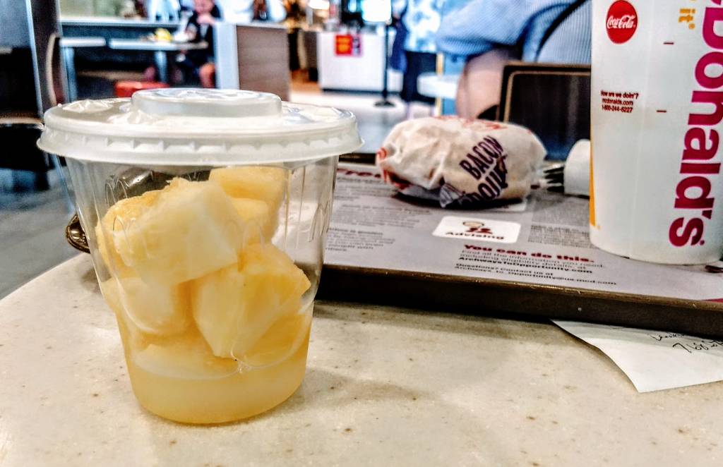 Copo transparente com pedaços de abacaxi e suco, ao lado de um sanduíche embrulhado e um copo de refrigerante em uma bandeja de fast food, com pessoas desfocadas ao fundo