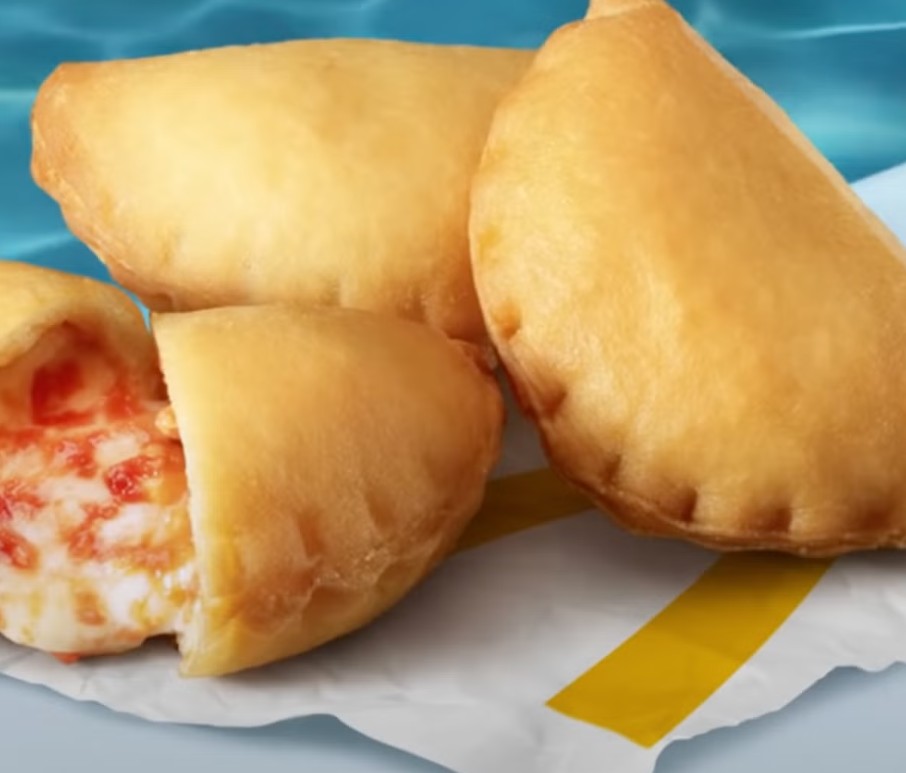 Três pasteizinhos fritos dourados sobre papel branco com faixa amarela, um deles cortado ao meio revelando recheio de queijo derretido e molho de tomate, com fundo azul ondulado
