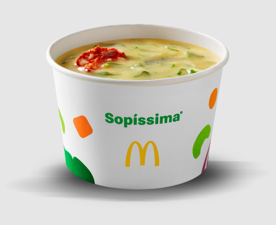 Copo branco de sopa Sopíssima do McDonald's, com caldo cremoso amarelo-esverdeado, pedaços de linguiça calabresa e vegetais. O copo tem o logo McDonald's e detalhes coloridos em verde, laranja e roxo