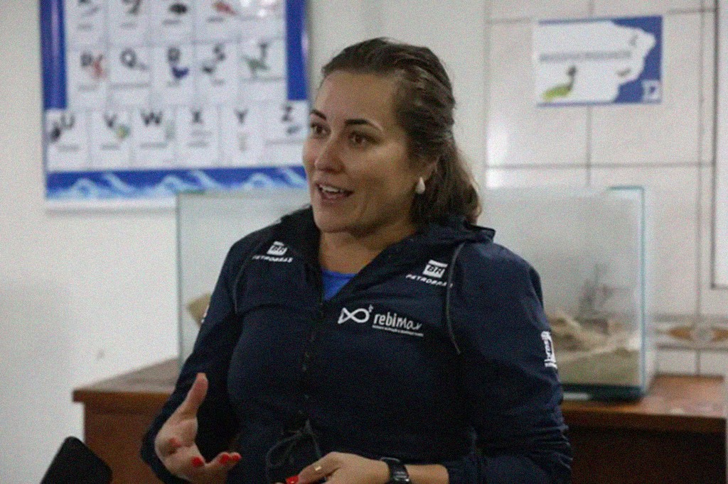 Mulher de cabelos castanhos presos, sorrindo e gesticulando, vestindo jaqueta azul-marinho com logotipos da Petrobras e Rebimar. Ela usa brincos de pérola e esmalte vermelho. Ao fundo, um quadro com letras e imagens, e um aquário sobre um móvel de madeira