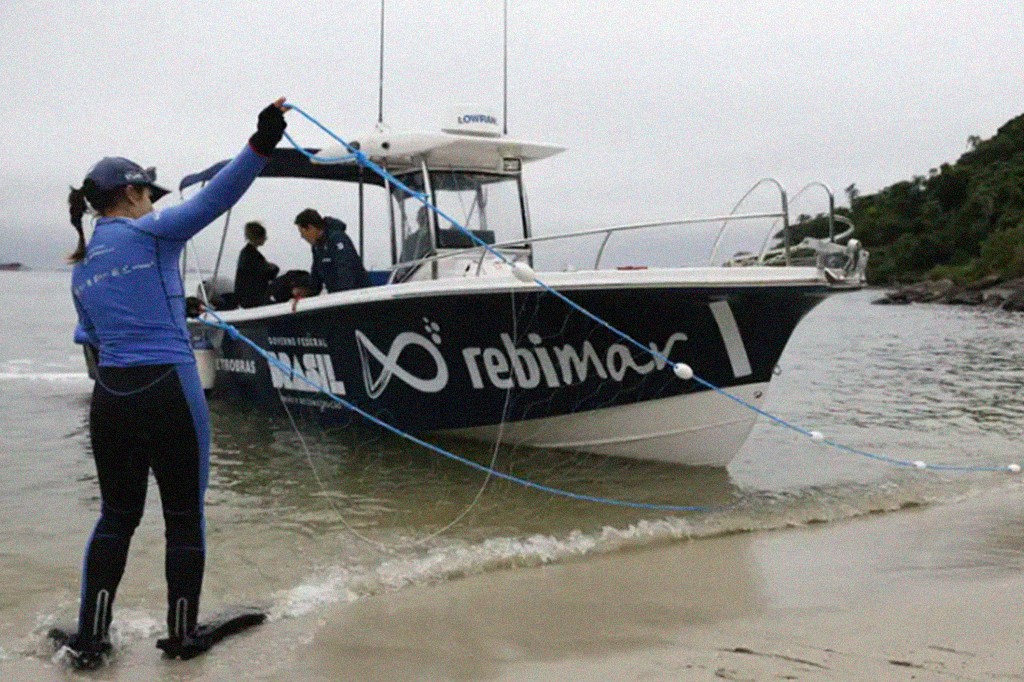 Mulher de roupa de mergulho azul e preta, com nadadeiras, puxa uma corda azul com boias brancas da água, em uma praia. Atrás dela, uma lancha branca e preta com o logo