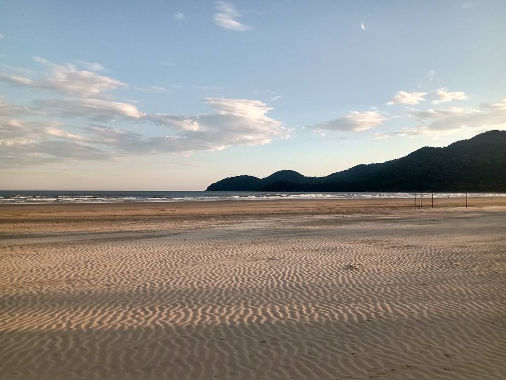 Praia deserta com areia clara e ondulada em primeiro plano, mar calmo ao centro e montanhas verdes escuras no horizonte sob um céu azul claro com nuvens esparsas