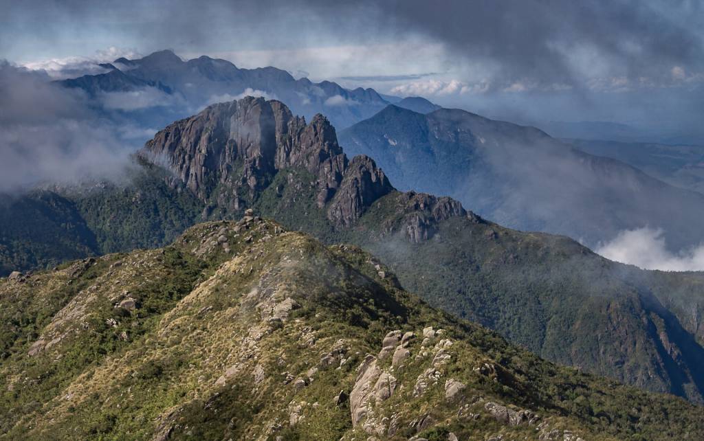Cadeia de montanhas rochosas e escarpadas, com vegetação verde e amarela, sob céu nublado e com nuvens baixas entre os picos