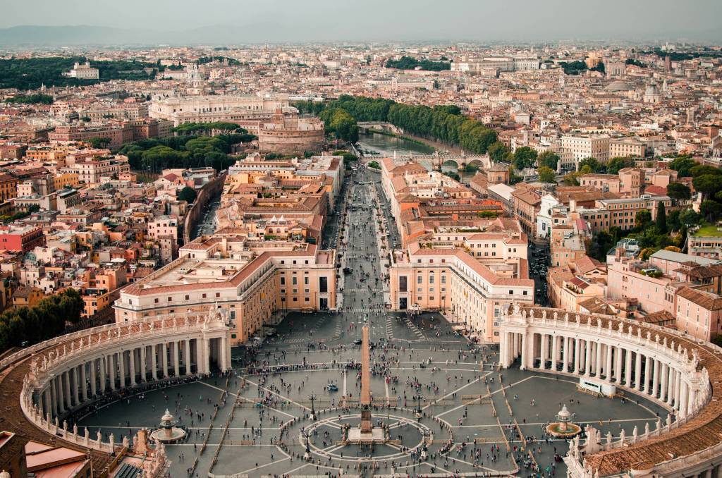 Vista aérea da Praça de São Pedro, no Vaticano, com milhares de pessoas reunidas. A praça oval é cercada por colunatas semicirculares e um obelisco central. Ao fundo, a cidade de Roma se estende com edifícios históricos e o rio Tibre