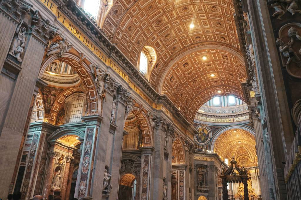 Interior da Basílica de São Pedro, com teto abobadado ricamente decorado em dourado e branco, arcos ornamentados com esculturas e colunas imponentes. A luz natural entra por janelas altas, iluminando a grandiosidade do local