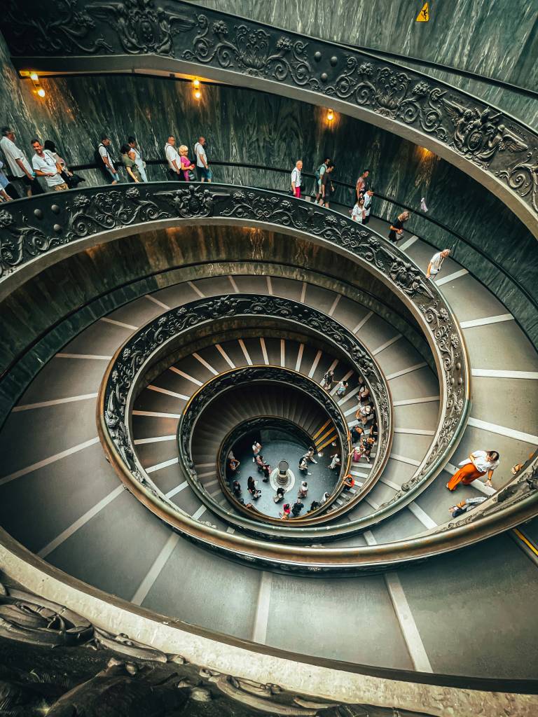 Vista aérea da escadaria em espiral de Giuseppe Momo no Museu do Vaticano, com dezenas de turistas descendo e subindo. A escada tem corrimãos de ferro forjado ornamentados e degraus cinzas com faixas brancas, e paredes de mármore escuro. No centro, um pilar branco e pessoas aglomeradas
