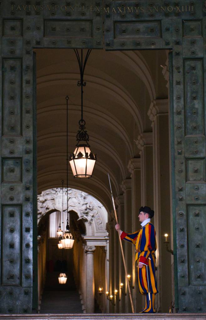 Guarda suíço jovem, de perfil, em uniforme listrado azul, vermelho e amarelo, segurando uma alabarda, em uma entrada de pedra com portas de bronze abertas e luminárias penduradas, com um corredor abobadado ao fundo