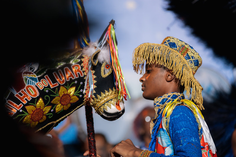 Jovem negro de perfil, com chapéu dourado de franjas e roupa azul e colorida, olhando para uma cabeça de cavalo de brinquedo preta, decorada com fitas coloridas, bordados de girassóis e a frase FILHO DO LUAR