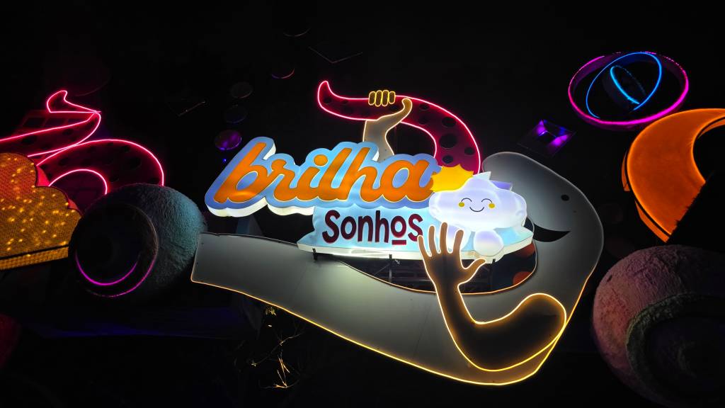 Letreiro neon colorido com a frase Brilha Sonhos em destaque. A palavra Brilha é laranja e azul, enquanto Sonhos é azul claro. Um braço neon amarelo segura uma nuvem sorridente, e um tentáculo rosa com bolinhas escuras aparece ao fundo, tudo em um ambiente escuro com outras luzes neon coloridas