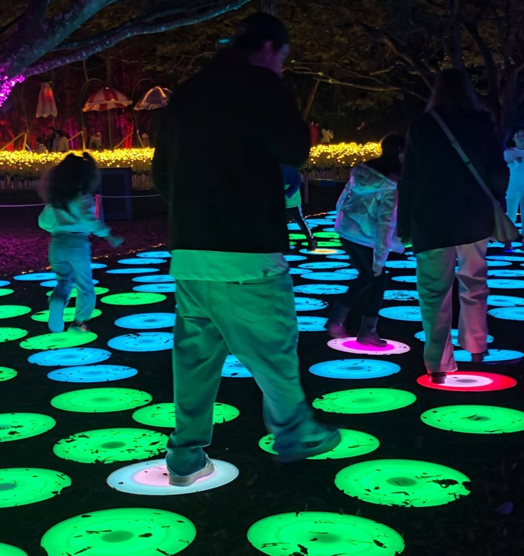 Pessoas caminham sobre discos luminosos coloridos no chão, em um ambiente noturno com árvores e luzes decorativas ao fundo. Os discos alternam entre verde, azul, rosa e branco, criando um caminho interativo.