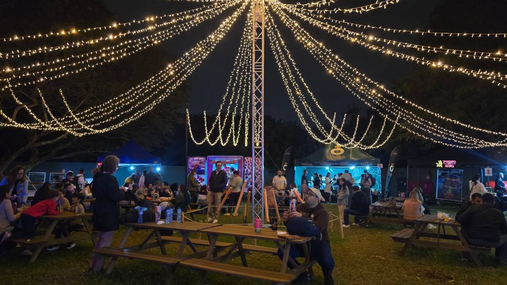 Pessoas em um evento noturno ao ar livre, sentadas em mesas de piquenique ou em pé, sob um céu escuro iluminado por muitas luzes de fada penduradas em zigue-zague. Há barracas de comida e bebida ao fundo, com algumas pessoas na fila. O ambiente é festivo e movimentado