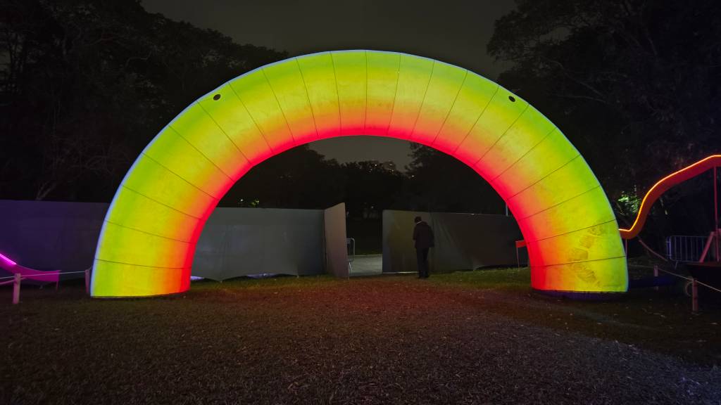 Um arco inflável gigante, iluminado em tons de verde, amarelo e vermelho, em um parque escuro à noite. Um homem de casaco escuro está de costas, caminhando sob o arco, que se destaca sobre a grama e árvores ao fundo