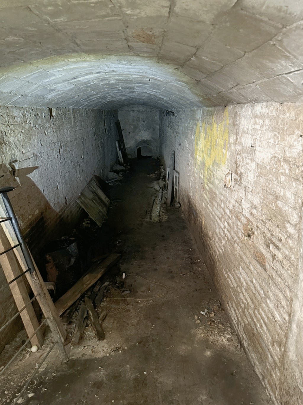 Corredor subterrâneo escuro e úmido com teto abobadado de tijolos. As paredes de tijolos brancos estão descascadas e manchadas, com lixo e tábuas de madeira espalhados pelo chão de terra batida. Uma mancha amarela vibrante se destaca na parede direita, enquanto o fundo do corredor é quase totalmente escuro, com uma pequena abertura arqueada visível