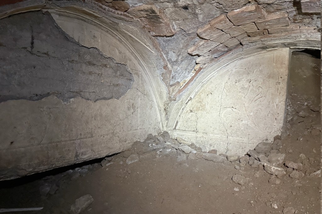Interior de uma estrutura antiga com teto abobadado de tijolos e paredes de argamassa desbotada. Há duas abóbadas com detalhes em relevo e parte do reboco descascado, revelando a estrutura interna. O chão é de terra com entulho de pedras e argamassa, sugerindo um local em ruínas ou em escavação