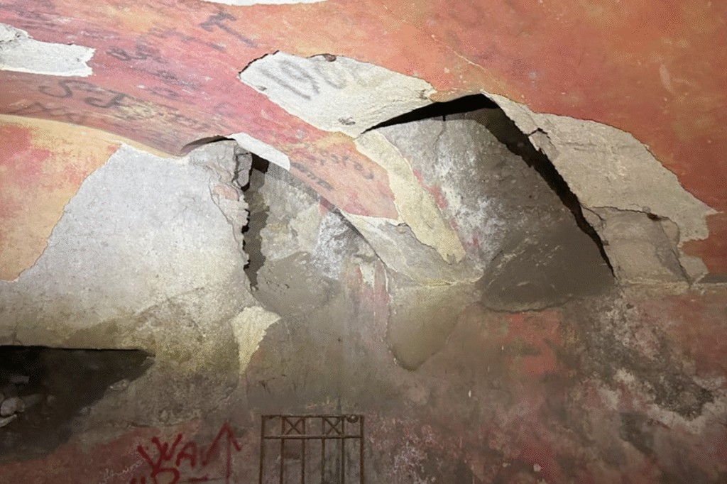 Teto abobadado de um túnel subterrâneo com a pintura vermelha descascando e revelando concreto e tijolos. Há pichações em preto e vermelho, e uma grade de metal enferrujada na parte inferior