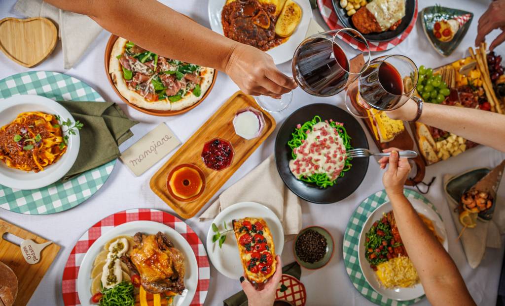 Mãos de várias pessoas brindam com taças de vinho tinto sobre uma mesa farta de pratos italianos, incluindo massas, pizza, frango assado, bruschettas, queijos e sobremesas, em uma toalha branca