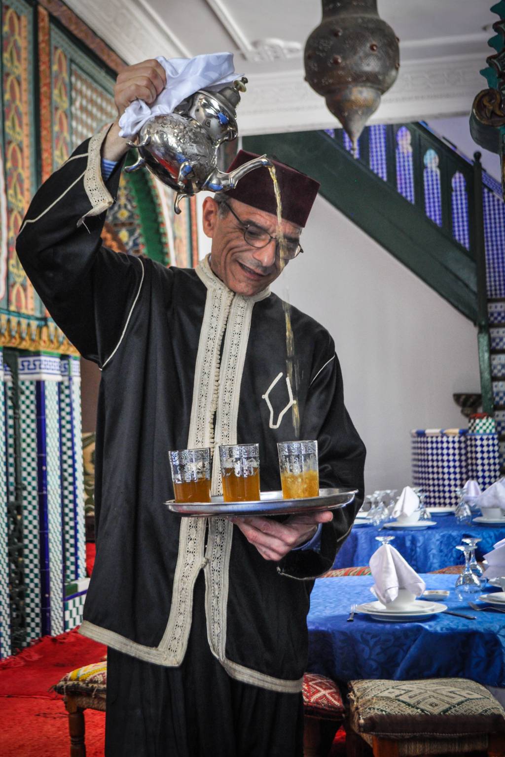Homem sorridente, de óculos e chapéu marrom, veste túnica preta com detalhes brancos, servindo chá de uma chaleira prateada em três copos de vidro sobre uma bandeja, em um ambiente com decoração árabe e mesas azuis