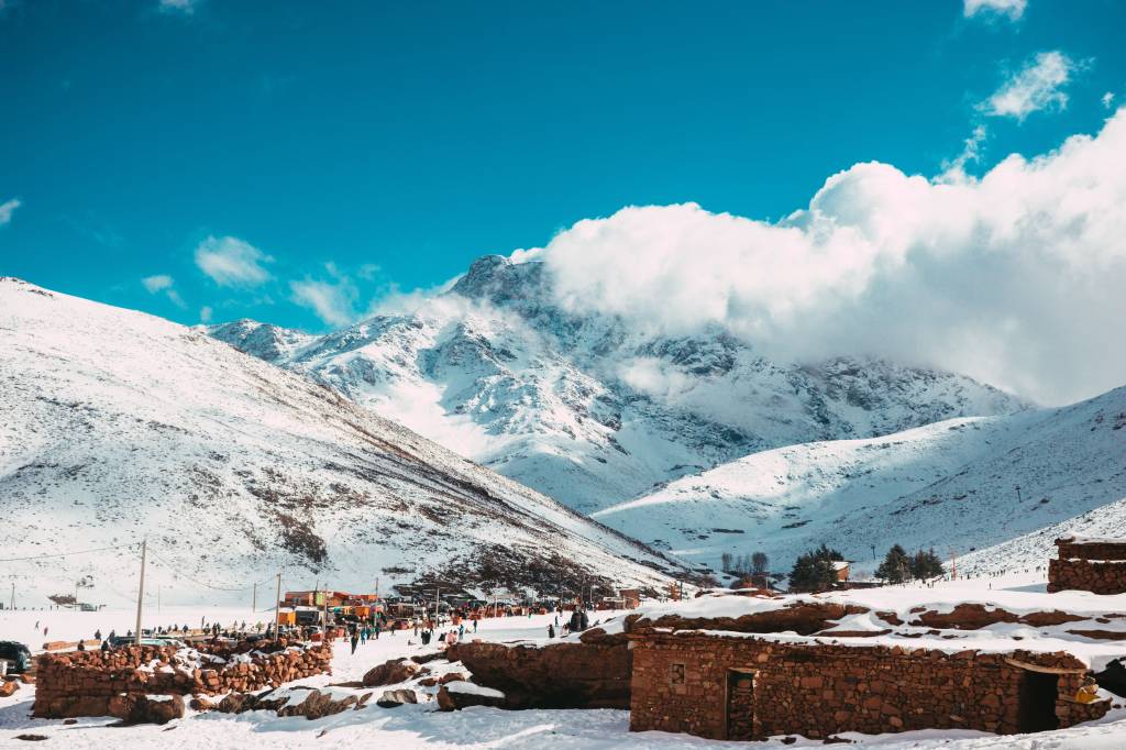 Montanhas nevadas sob um céu azul vibrante com nuvens brancas. No vale, um vilarejo com casas de pedra e muitas pessoas caminhando na neve, algumas perto de barracas coloridas. O cenário é de inverno, com picos cobertos de neve e um clima frio.
