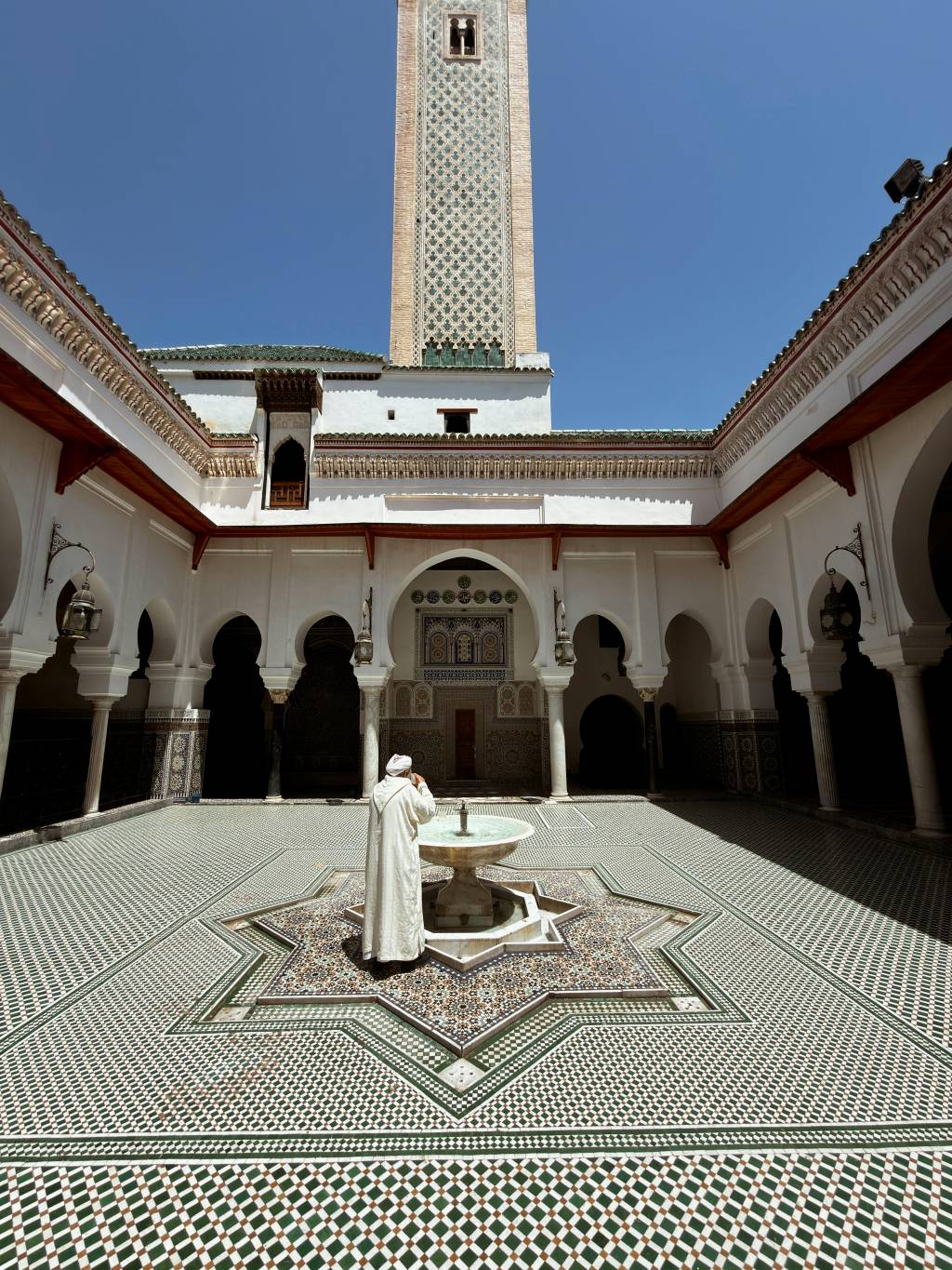 Homem de túnica branca e turbante, de costas, em pátio de mesquita com fonte central. O chão tem mosaicos verdes e brancos, e ao fundo ergue-se um minarete decorado sob céu azul