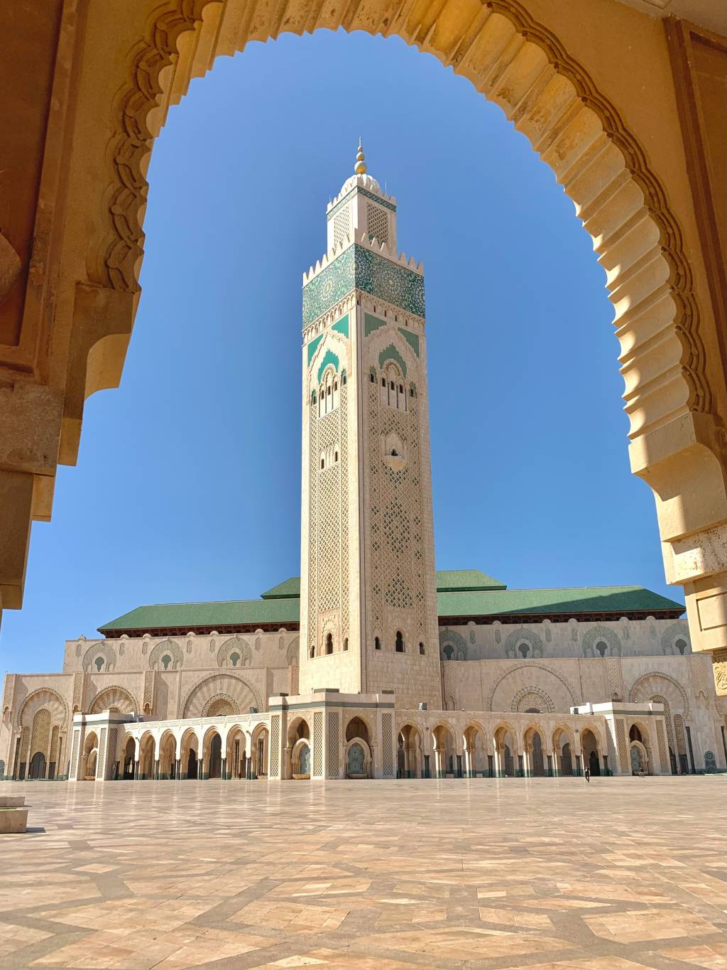 Minarete da Mesquita Hassan II em Casablanca, Marrocos, visto através de um arco ornamentado. O minarete alto e bege tem detalhes em mosaico verde e branco, com um telhado verde da mesquita ao fundo. O céu é azul claro e o pátio vazio tem piso de pedra com padrões geométricos