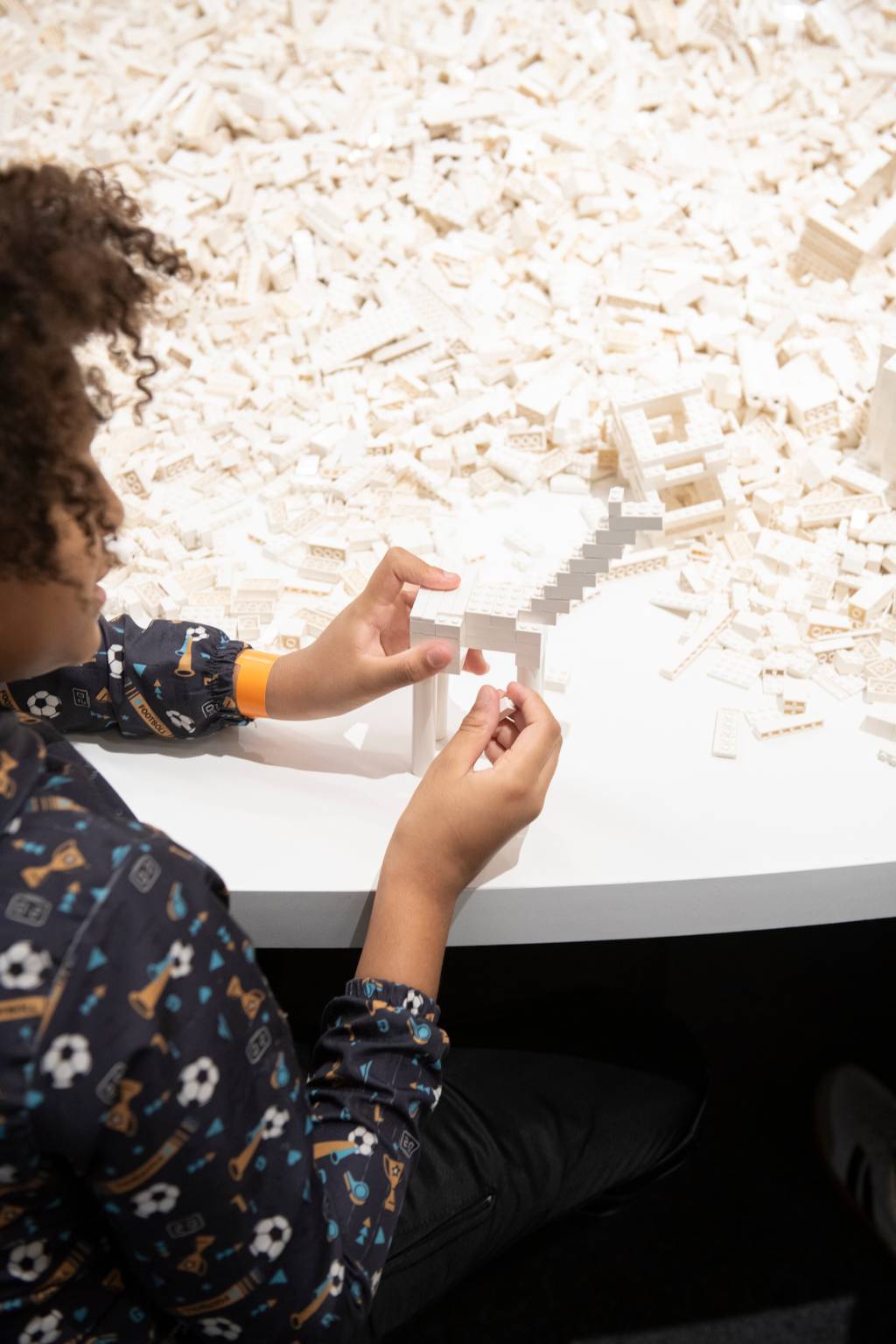 Criança com cabelo cacheado e camisa estampada de futebol, sentada em uma mesa branca, constrói uma estrutura com peças de Lego brancas. Ao fundo, um monte de peças brancas.