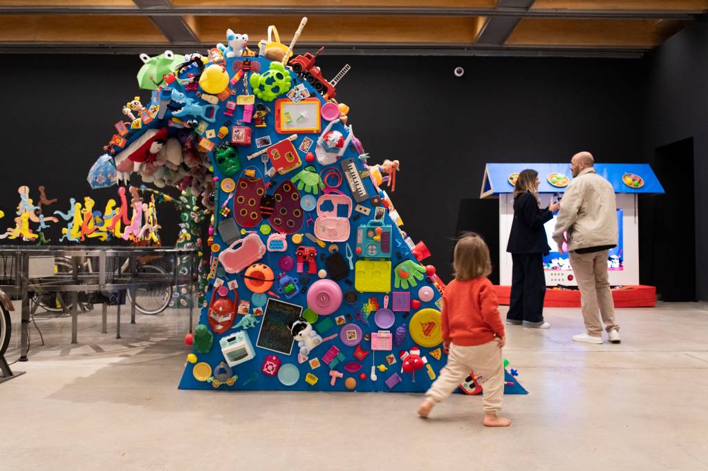 Uma criança de cabelo loiro e blusa laranja caminha descalça em direção a uma escultura azul triangular coberta por centenas de brinquedos coloridos. Ao fundo, um casal conversa perto de uma casinha de brinquedo azul e branca. O ambiente é um museu com paredes escuras e piso claro