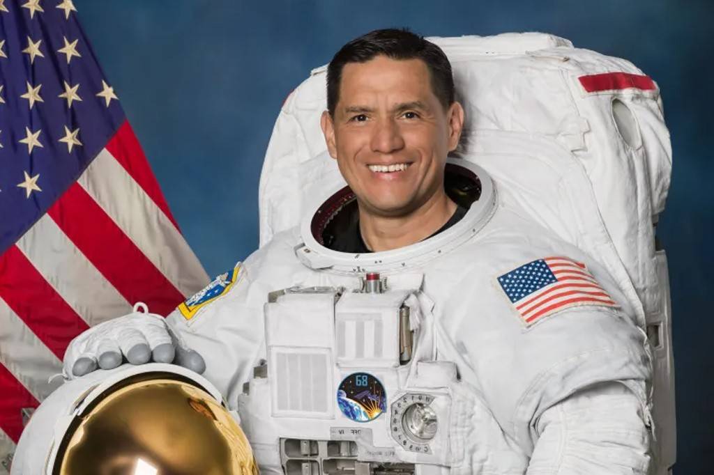 Astronauta sorridente, de pele morena e cabelo escuro, vestindo um traje espacial branco com a bandeira dos EUA no braço, posa com um capacete dourado na mão, em frente a uma bandeira americana e fundo azul