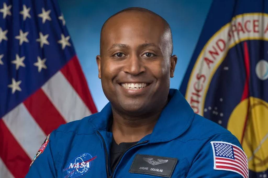 Homem negro careca sorrindo, vestindo uniforme azul da NASA, com bandeira dos EUA e logotipo da agência espacial ao fundo