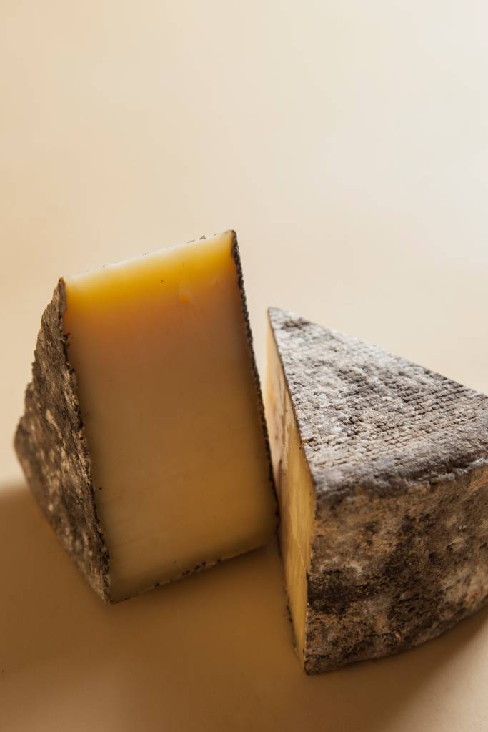 Dois pedaços de queijo artesanal, um triangular e outro retangular, com casca escura e rugosa, e interior amarelo claro, sobre fundo bege