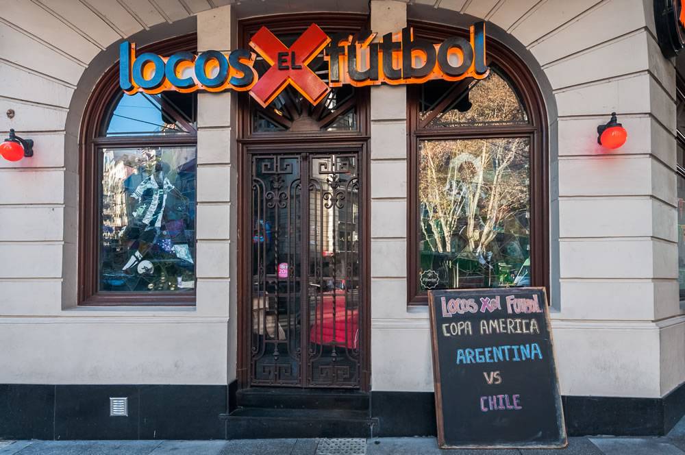 Fachada de um bar chamado Locos x el Fútbol, com letreiro azul e laranja. A vitrine exibe uma camisa de futebol listrada em preto e branco. Uma lousa na calçada anuncia Copa América Argentina vs Chile