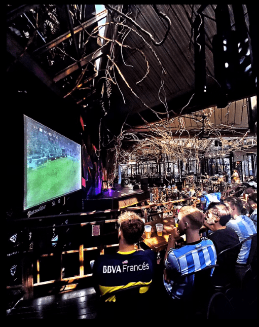 Pessoas assistem a um jogo de futebol em um telão, sentadas em um bar com decoração rústica de galhos secos no teto. Dois homens em primeiro plano vestem camisetas de times, um azul e amarelo, outro azul e branco, com copos de cerveja na mesa. O ambiente é escuro, com iluminação focada no telão e na decoração.