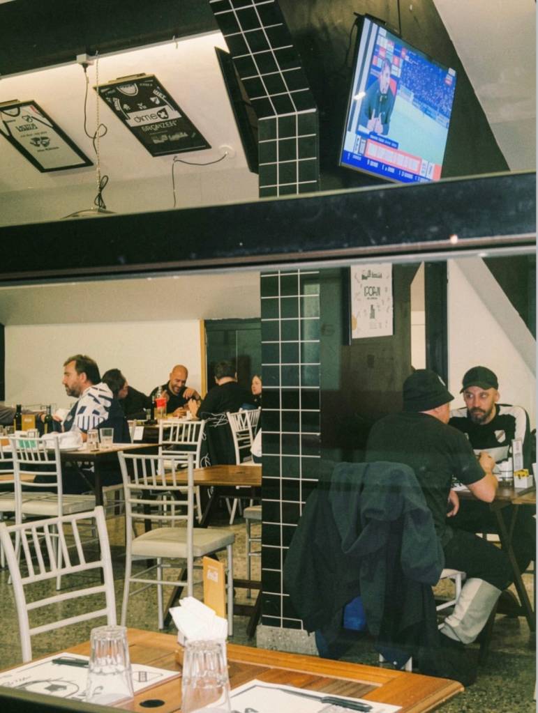 Pessoas sentadas em mesas de madeira em um restaurante com paredes pretas e brancas, assistindo a um jogo de futebol em uma TV suspensa