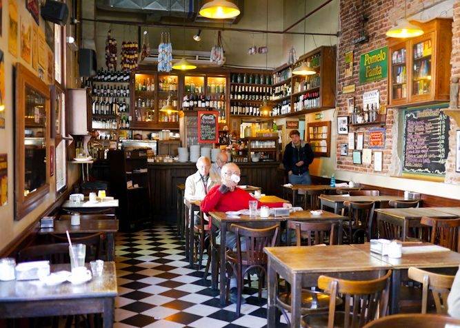 Interior de um café ou bar antigo com piso xadrez preto e branco, mesas e cadeiras de madeira escura. Ao fundo, um balcão com prateleiras cheias de garrafas e um homem de camisa azul. Dois homens idosos sentam-se em uma mesa no centro, um de camisa vermelha e outro de casaco claro. As paredes são de tijolos e decoradas com quadros e cartazes. A iluminação é amarela e aconchegante