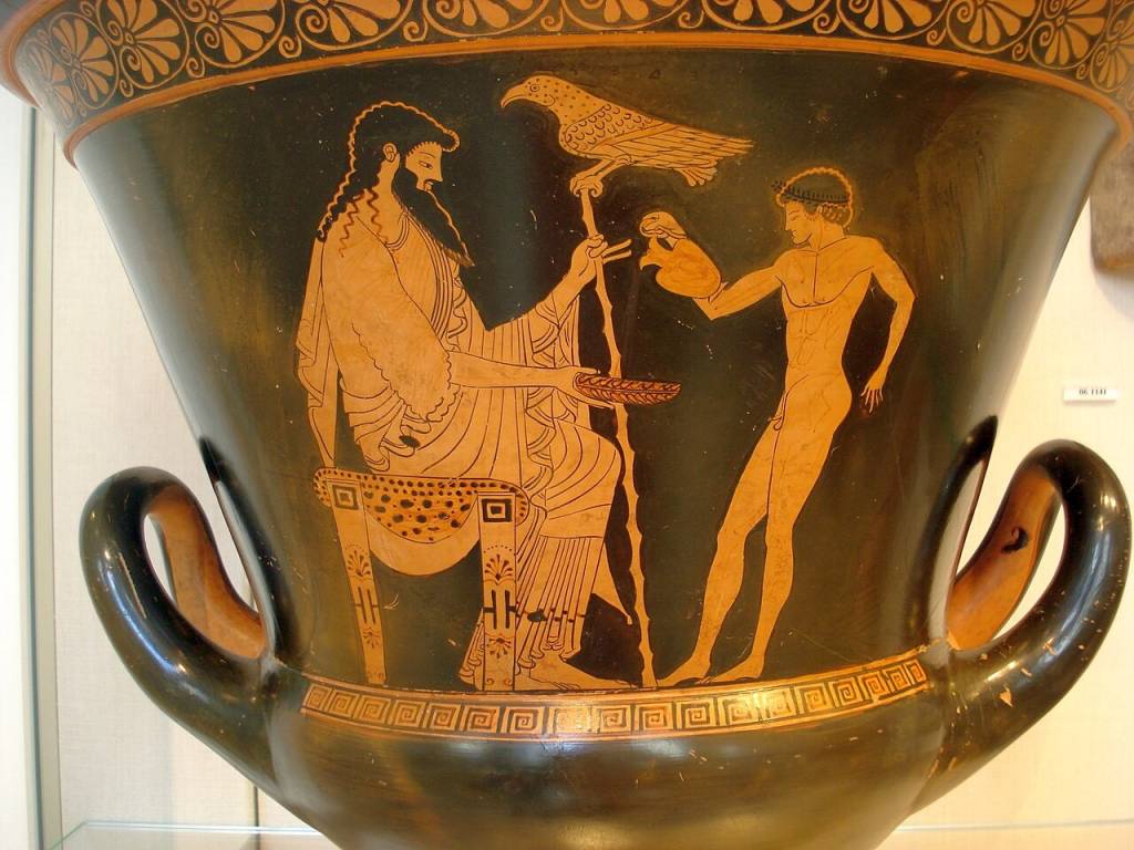 Pintura em vaso grego antigo, com fundo preto e figuras em terracota. Um homem barbudo e vestido senta-se à esquerda, segurando um cajado. Um pássaro pousa no cajado. À direita, um jovem nu oferece um objeto ao homem sentado. Alças laterais e padrões geométricos adornam o vaso