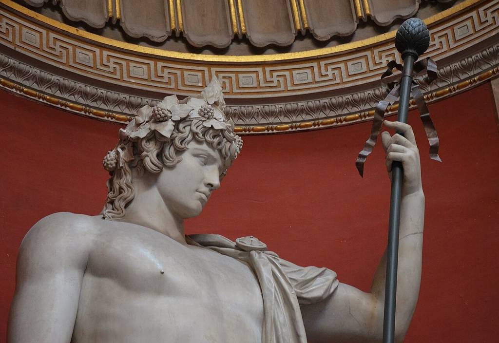 Estátua de mármore de um homem jovem, com coroa de folhas e uvas, segurando um bastão com uma pinha na ponta e fitas, contra uma parede vermelha e teto dourado com padrões geométricos