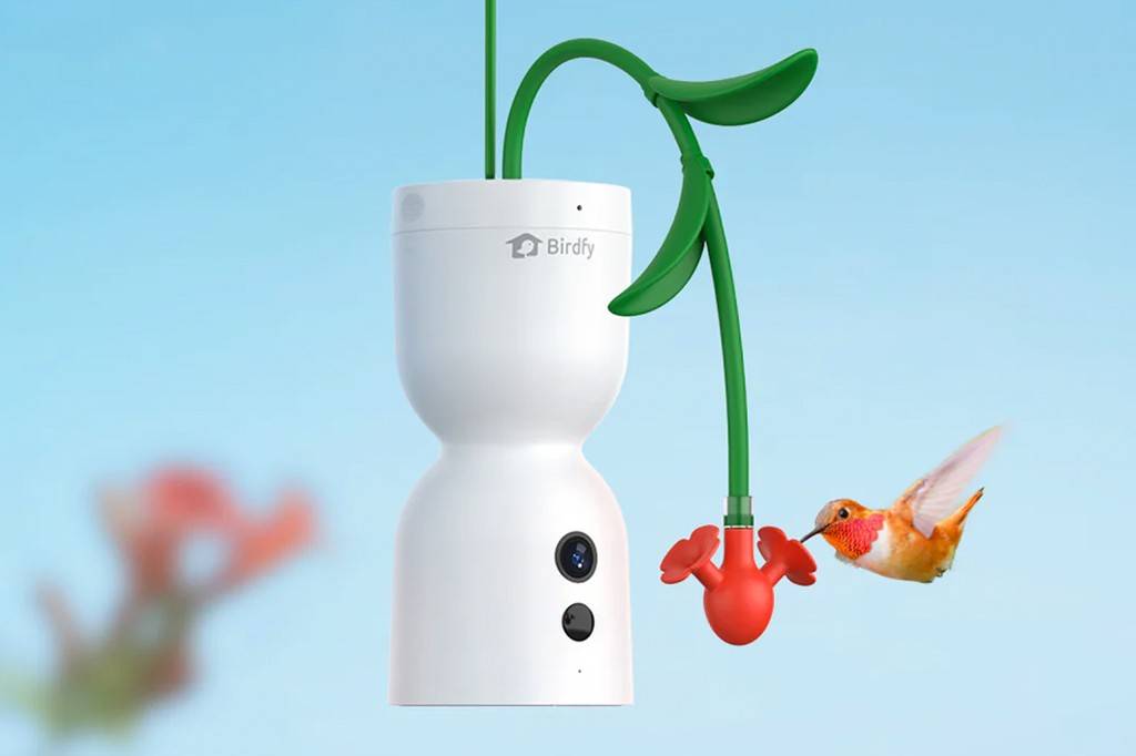 Câmera de vigilância Birdfy branca em formato de ampulheta, com um braço verde em forma de folha e um alimentador vermelho de beija-flor, enquanto um beija-flor laranja se aproxima para beber néctar. Fundo azul claro.