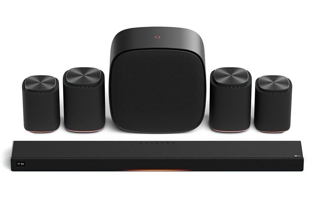 Sistema de som surround preto com soundbar retangular na frente, subwoofer central e quatro caixas de som cilíndricas ao redor, todos com detalhes em cobre na base.
