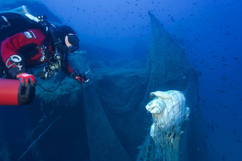 Mergulhador em roupa vermelha e preta observa uma tartaruga marinha presa em uma rede de pesca fantasma no fundo do oceano azul escuro, com pequenos peixes ao redor